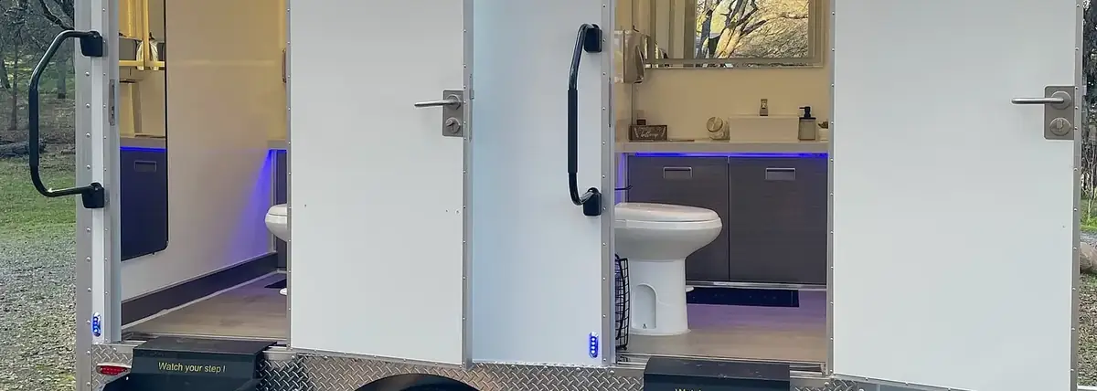 Two-stall restroom trailer with lit interior and steps on a gravel lot