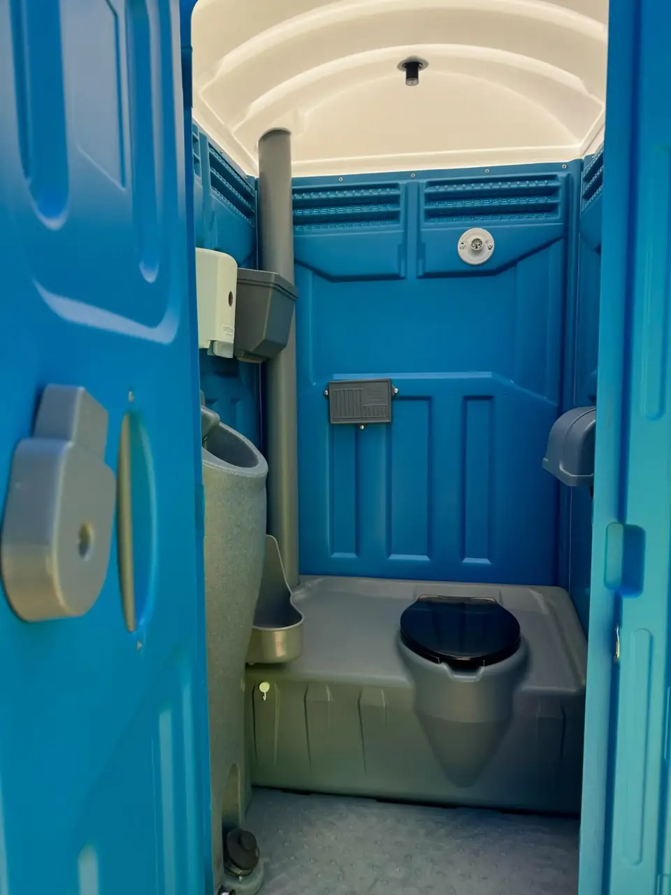 Inside a blue standard porta potty showing the toilet seat, wall urinal and toilet paper holder